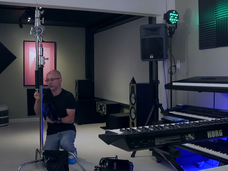 Wind, Sand, and Uneven Concrete: How to Choose a Light Stand for Location Shoots 1 A professional videographer adjusting a heavy-duty silver C-stand light stand in a studio setup with musical equipment.